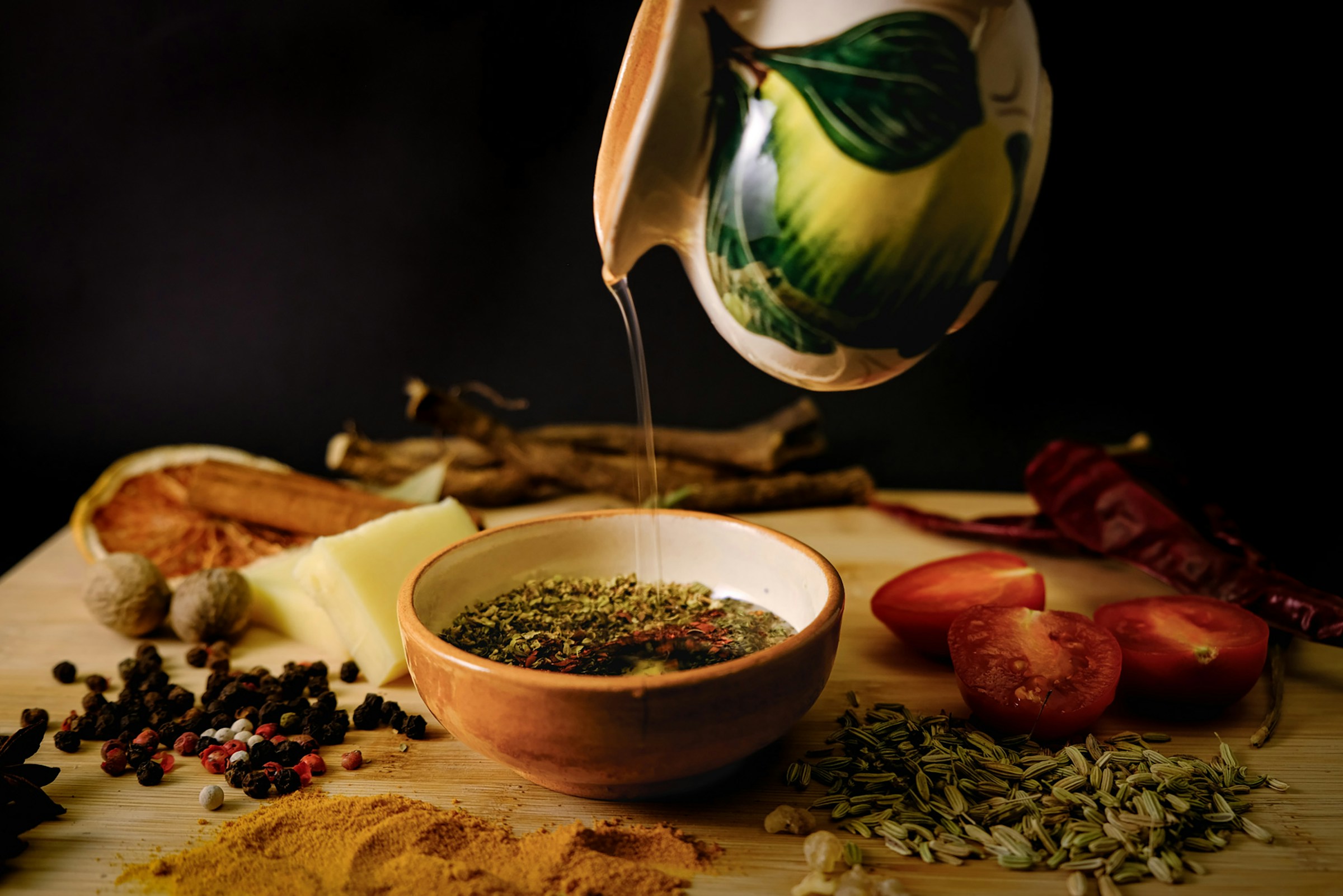 Olive oil pouring from a ceramic vessel into a bowl of herbs
